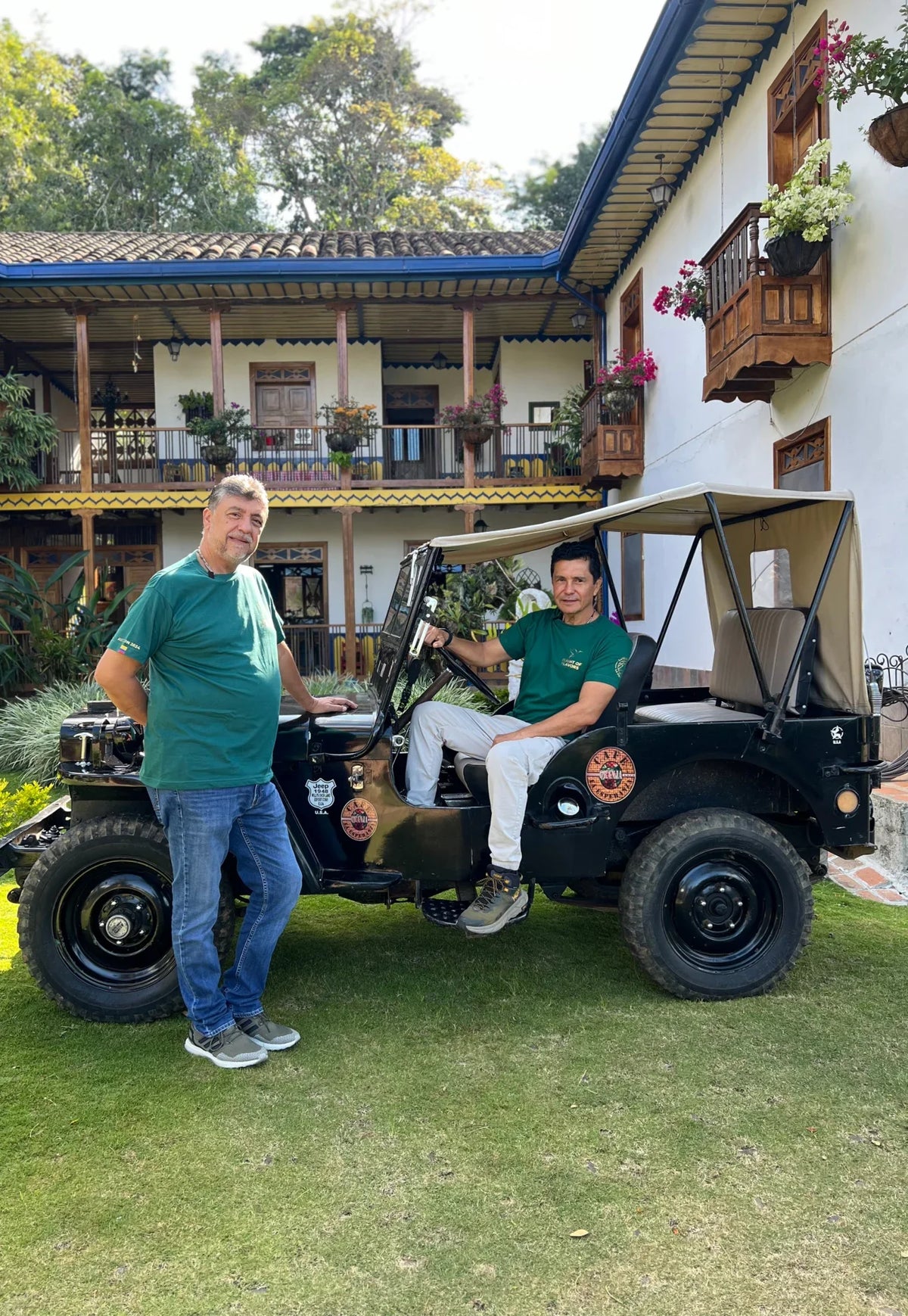 Dos hombres junto a un jeep frente a una casa con corredores y balcones de madera.