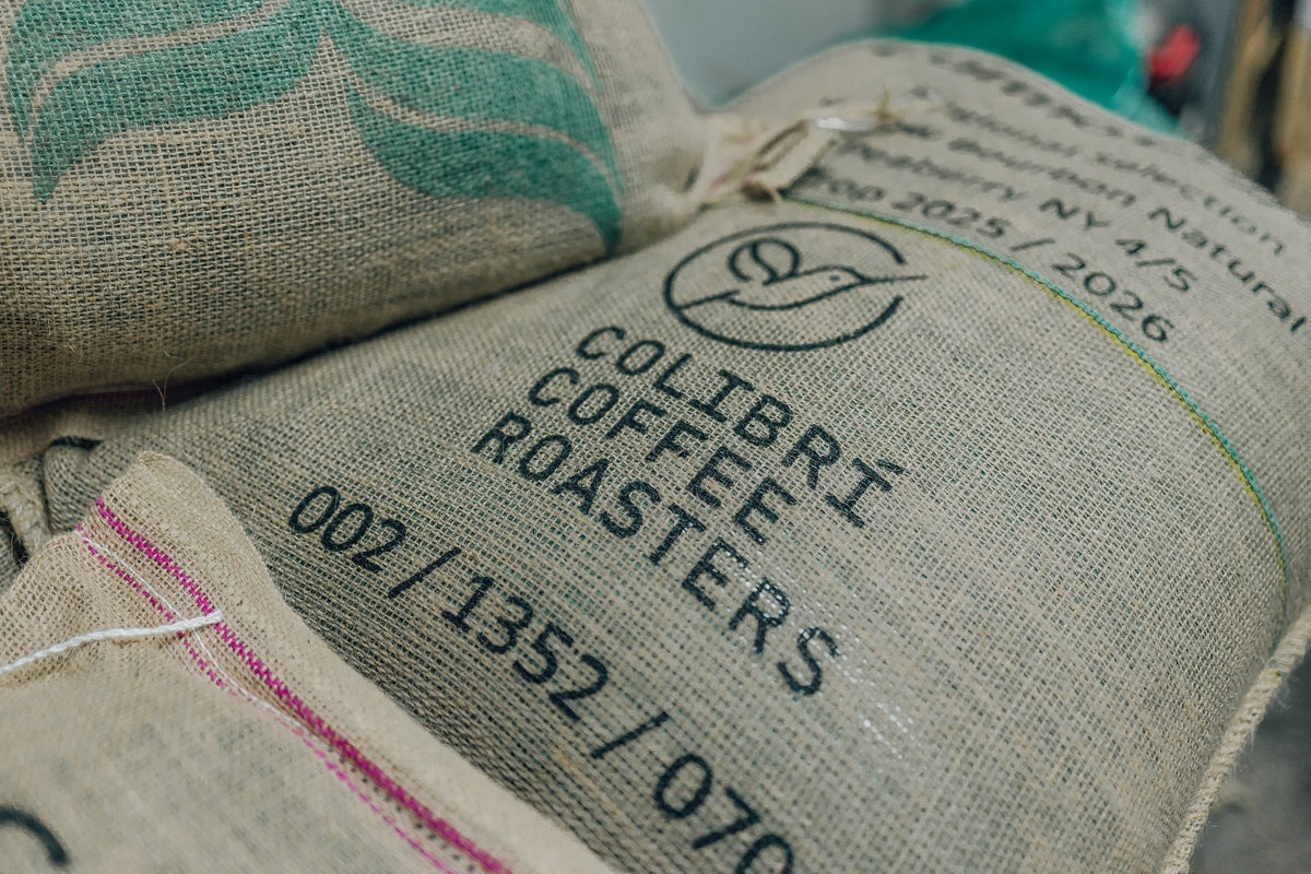 Sacos de yute apilados, marcados con Colibrí Coffee Roasters.